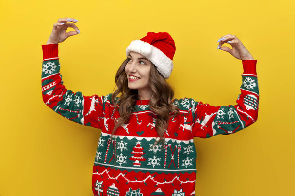 girl in Christmas sweater and santa claus hat holds empty hands on yellow isolated background, woman in New Year's clothes decorates and raises her hands - Photo, Image