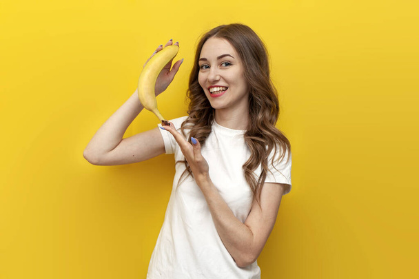 mladá roztomilá dívka ukazuje banán a usmívá se na žlutém izolovaném pozadí, žena drží ovoce a raduje - Fotografie, Obrázek