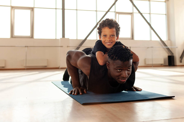 Afrikkalainen amerikkalainen mies poikansa kanssa menee urheiluun kuntosalilla, isä tekee punnerruksia lattialta poikansa kanssa ja opettaa terveellistä elämäntapaa. - Valokuva, kuva