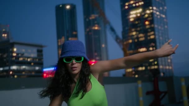 ナイトシティで踊るクールガールダンサー. 長いカーリー髪の黒い少女はリズム的に動きます. トレンディなバケツカップと明るい緑色のメガネでセクシーなファッショナブルなダンサーが感覚的に都市中心部を移動します - 映像、動画