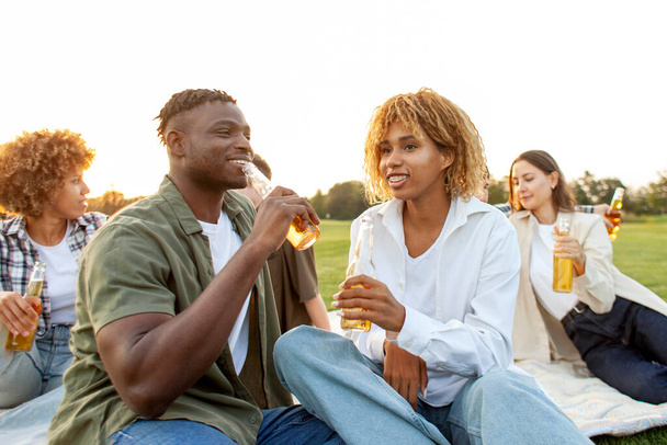 african american couple holding bottle of beer and talking in park outdoors, man and woman drinking alcohol and smiling at picnic with friends, group of people relaxing and celebrating - Photo, Image