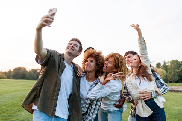 группа мультирасовых молодых людей делает селфи на смартфоне и фотографирует в парке, межрасовые студенты общаются через видеосвязь онлайн на открытом воздухе - Фото, изображение