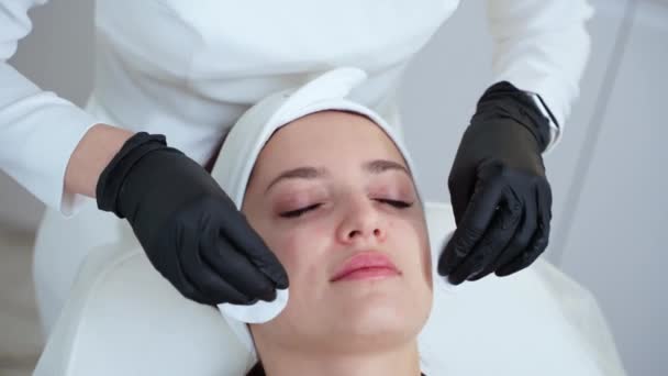 donna adulta è rilassante nel salone spa durante il trattamento di cura della pelle, estetista sta asciugando la pelle del viso della paziente femminile da rilievi con lozione idratante, primo piano del viso - Filmati, video