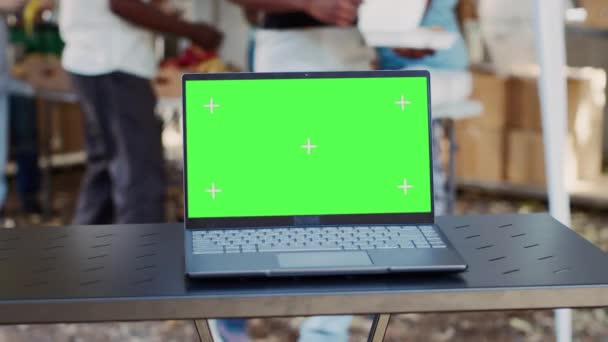Крупним планом електронний ноутбук з ізольованим зеленим екраном сидить на столі, тоді як благодійні працівники допомагають менш привілейованим. Детальний перегляд мінікомп'ютера з порожнім шаблоном макета копій. - Кадри, відео