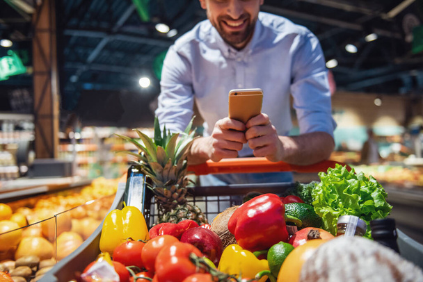 Обрізане зображення красивого чоловіка, який використовує мобільний телефон і посміхається під час покупки в супермаркеті
 - Фото, зображення