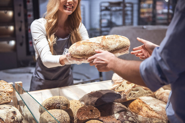 Bij de supermarkt. Bijgesneden afbeelding van mooie jonge vrouwelijke werknemer een brood aan de klant bieden en glimlachen - Foto, afbeelding