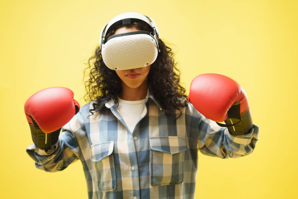 Πορτρέτο μιας νεαρής γυναίκας που φοράει γυαλιά εικονικής πραγματικότητας και γάντια πυγμαχίας κρατώντας τα χέρια της σε θέση φρουράς, απολαμβάνοντας το παιχνίδι πυγμαχίας, gamer και χόμπι σε κίτρινο φόντο.  - Φωτογραφία, εικόνα