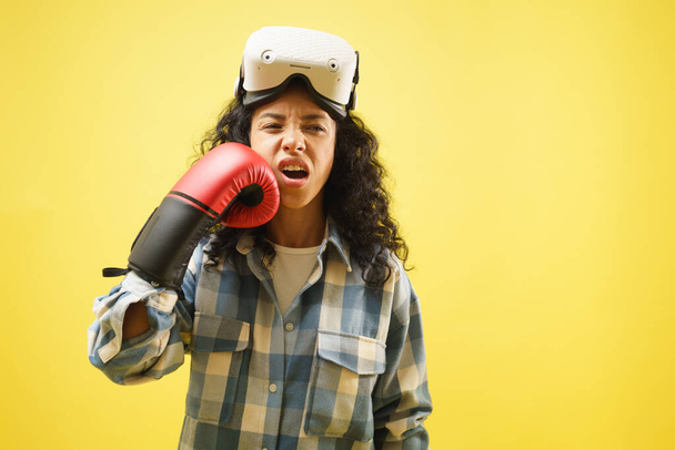 Fille portant VR masque et gant de boxe agissant avec punch elle-même sur son visage sur le fond jaune. - Photo, image