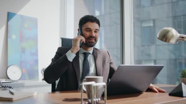 Mosolygós menedzser hívja a modern irodai asztalnál ülő ügyfelet. Boldog pozitív üzletember, kellemes telefonbeszélgetést folytat a munkahelyén. Vidám szakállas vállalati dolgozó beszél telefon a munkaszünetben - Felvétel, videó