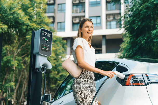 Young woman travel with EV electric car charging in green sustainable city outdoor garden in summer. Urban sustainability lifestyle by green clean rechargeable energy of electric BEV vehicle innards - Photo, Image