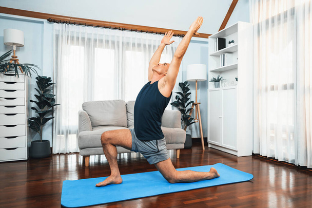 Gelukkig actieve senior man in sportkleding doet yoga houding op trainingsmat thuis. Gezonde seniorenlevensstijl met yoga-oefening. Klok - Foto, afbeelding