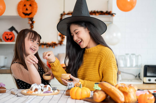 Een gelukkig en vrolijk jong Aziatisch meisje en haar moeder in Halloween kostuums lachen, hebben plezier, genieten van het versieren van Halloween cupcakes in de keuken samen. Spookachtig seizoen, familietijd - Foto, afbeelding