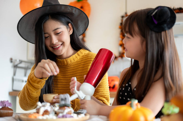 Een vriendelijke en mooie jonge Aziatische moeder met een heksenhoed maakt graag Halloween cupcakes met haar jonge dochter in de keuken, om thuis Halloween te vieren. Halloween griezelig seizoen concept - Foto, afbeelding