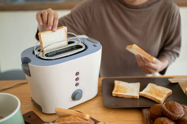 Close-up image of a beautiful young Asian woman is making tasty toasts with a toaster in the kitchen, preparing her breakfast. Cooking concept - Photo, Image