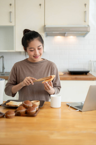 Una bella e felice giovane asiatica sta spargendo marmellata di fragole su un toast, preparando la sua colazione in cucina. Concetto di cucina domestica - Foto, immagini