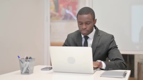 Afrikanischer Geschäftsmann blickt auf Kamera, während er im Büro am Laptop arbeitet - Filmmaterial, Video