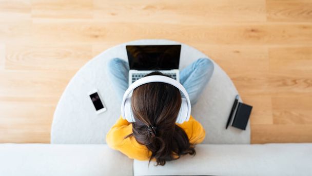 Κάτοψη της νεαρής Ασιάτισσας που κάθεται στο πάτωμα και εργάζεται σε φορητό υπολογιστή με βιβλίο, στυλό και κινητό smartphone, φορώντας ασύρματα ακουστικά, ακούγοντας μουσική - Φωτογραφία, εικόνα