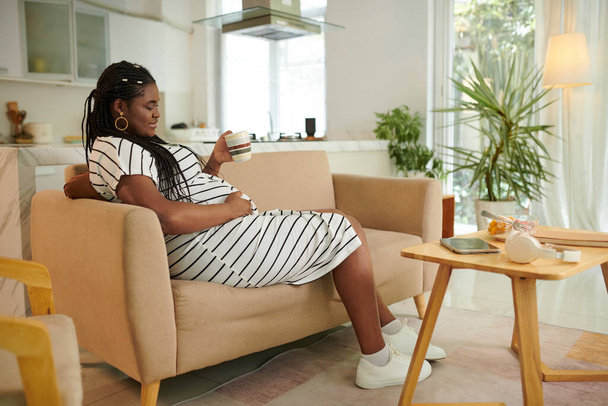 Mujer embarazada sonriente descansando en el sofá y disfrutando de una taza de té - Foto, Imagen