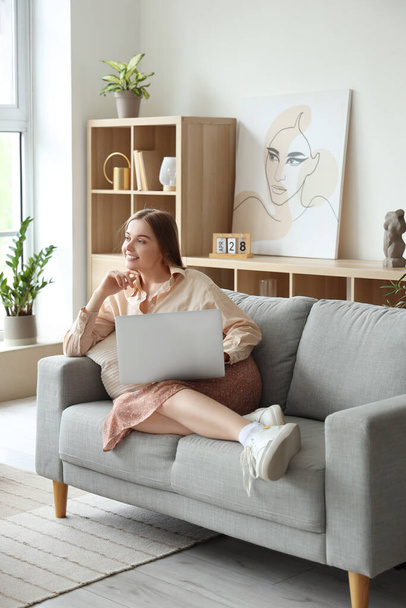 Mujer bastante joven sentada en un sofá gris y usando un portátil moderno en la sala de estar ligera - Foto, imagen