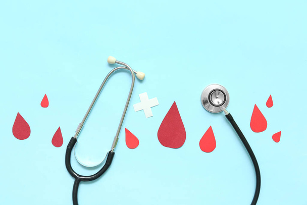 Stetoskop z papierowymi kroplami krwi i krzyżem na niebieskim tle. Światowy Dzień Dawcy - Zdjęcie, obraz