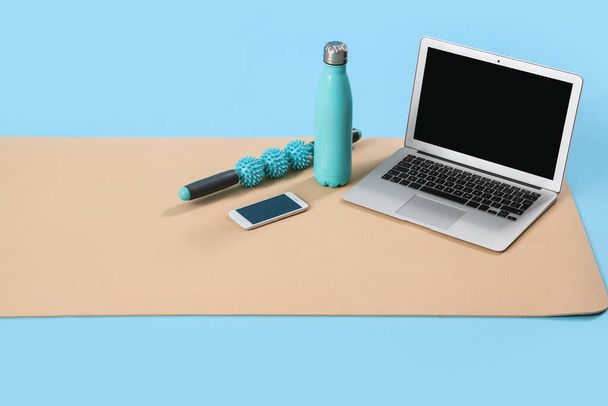 Фітнес килимок з ноутбуком, мобільним телефоном, пляшкою та масажним роликом на синьому фоні - Фото, зображення