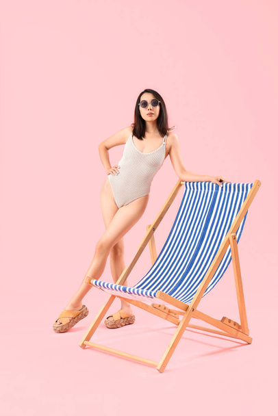Beautiful Asian woman in swimsuit with deck chair on pink background - Photo, Image