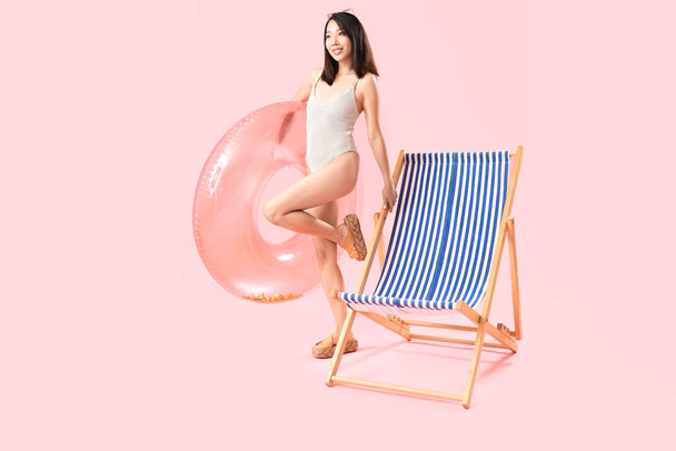 Beautiful Asian woman with swim ring and deck chair on pink background - Photo, Image