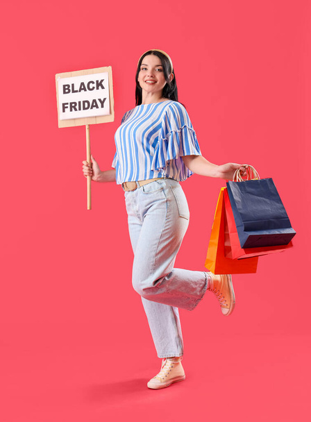 Hermosa mujer joven sosteniendo bolsas de compras y firmar con texto VIERNES NEGRO sobre fondo rojo - Foto, imagen