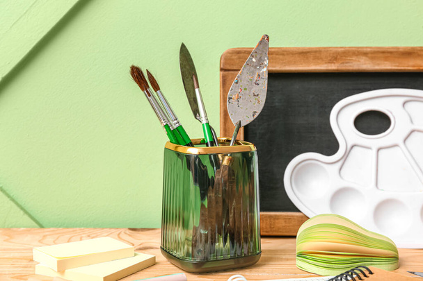 Holder with different painter's supplies and chalkboard on wooden desk, closeup - Photo, Image
