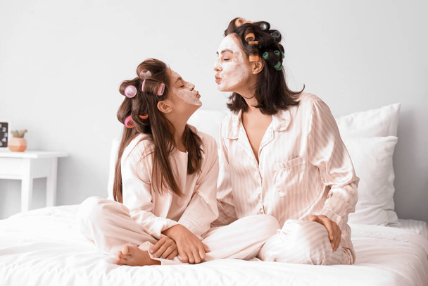Маленькая девочка и ее мать с бигуди для волос наносят маску для лица в спальне - Фото, изображение