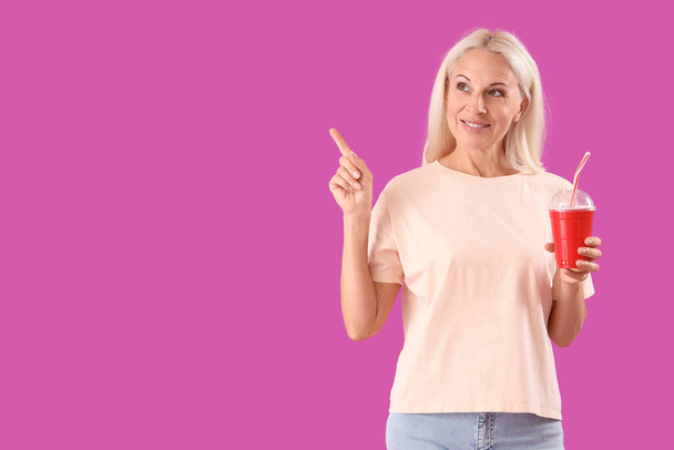volwassen vrouw met kopje frisdrank wijzend naar iets op paarse achtergrond - Foto, afbeelding