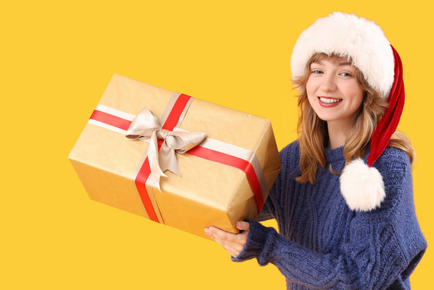Mujer joven en sombrero de Santa con regalo de Navidad sobre fondo amarillo - Foto, Imagen