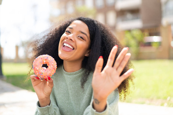 Jeune femme afro-américaine tenant un beignet à l'extérieur saluant avec la main avec expression heureuse - Photo, image
