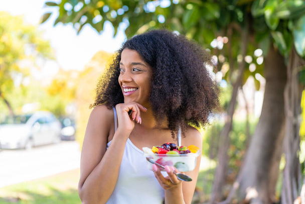 Młoda Afroamerykanka trzyma miskę owoców na świeżym powietrzu myśląc o pomyśle i patrząc w bok - Zdjęcie, obraz