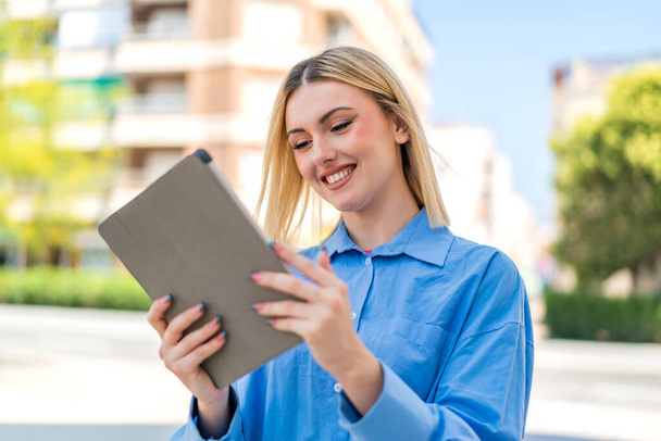 Jeune jolie femme blonde à l'extérieur tenant une tablette avec une expression heureuse - Photo, image