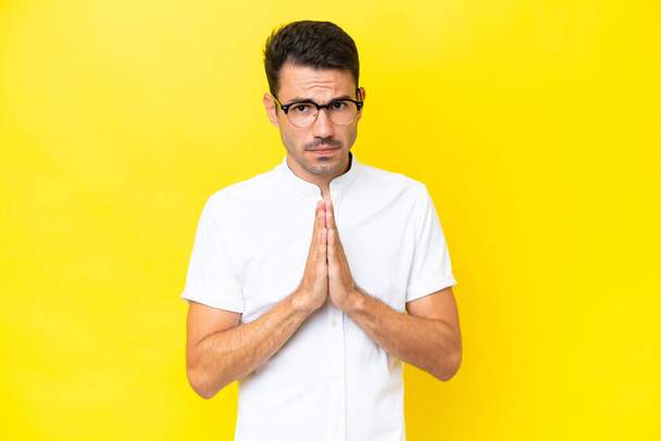 Young handsome man over isolated yellow background keeps palm together. Person asks for something - Photo, Image
