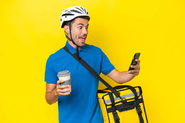 Fiatal jóképű férfi termikus hátizsák felett elszigetelt sárga háttér kezében kávét, hogy vigye el, és egy mobil - Fotó, kép