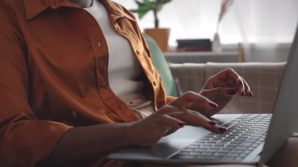 Inclinación hacia arriba de la joven mujer afroamericana freelance en auriculares inalámbricos que trabajan en el ordenador portátil en la oficina en casa - Imágenes, Vídeo