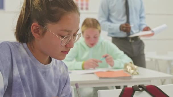 Borst omhoog van Kaukasische tiener meisje in bril doen test tijdens de les in helder klaslokaal met zwarte mannelijke leraar helpen andere meisje op de achtergrond - Video