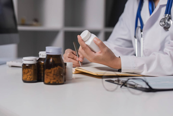 Азіатська жінка-лікар gp терапевт тримає пляшку таблеток в руці, пишучи медичний рецепт, сидячи на робочому столі, прописуючи концепцію аптечної медицини. Перегляд крупним планом. - Фото, зображення
