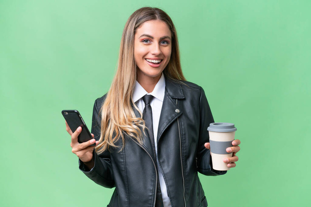 Бізнес досить Уругвайська жінка вдягнена в велосипедну куртку над ізольованим фоном утримуючи каву, щоб забрати її і мобільний телефон - Фото, зображення