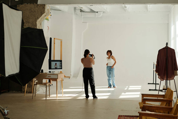 Вид спереду молодого професійного фотографа з фотокамерою, що стоїть перед жіночою моделлю моди в повсякденному одязі, позуючи на білій стіні - Фото, зображення