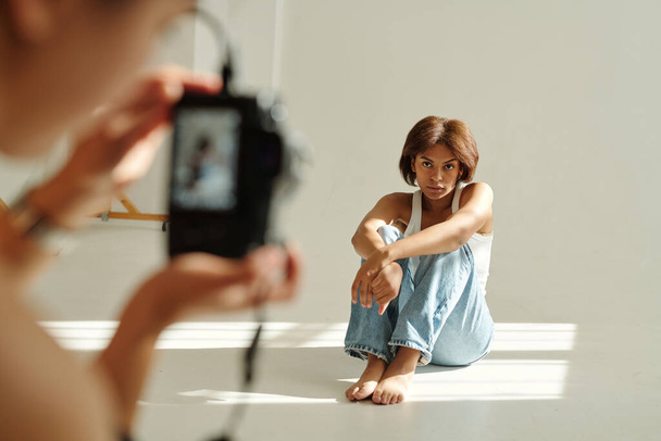 Jeune jolie mannequin de mode afro-américaine en casual posant pour appareil photo tout en étant assis devant le photographe pendant la séance photo - Photo, image