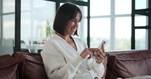 Tevreden vrouw zit op de woonkamer bank, surfen op haar telefoon met een glimlach van geluk. Haar ontspannen houding en vrolijke expressie weerspiegelen de eenvoudige vreugde van het verkennen van digitale inhoud. - Video