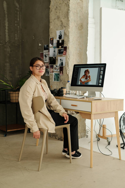 Genç profesyonel fotoğrafçı iş yerinde bilgisayar monitörüyle otururken kameraya bakıyor. Moda modelinin görüntüleri ekranda. - Fotoğraf, Görsel