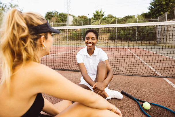 Dos mujeres hermosas jóvenes teniendo una conversación sentadas en una cancha de tenis al lado de la red. Dos jugadores de tenis amateurs tomando un descanso durante un partido de tenis. - Foto, Imagen