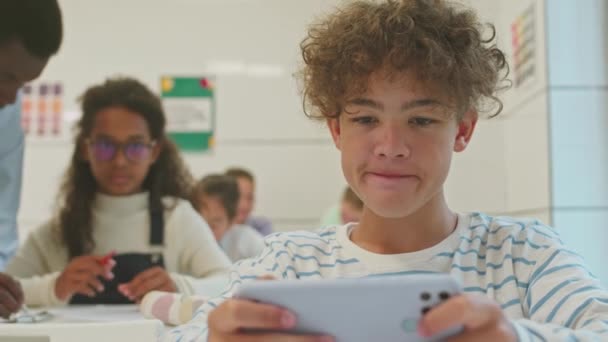 Cintura hacia arriba del estudiante de sexto grado Biracial masculino jugando juegos en el teléfono inteligente durante la lección - Imágenes, Vídeo