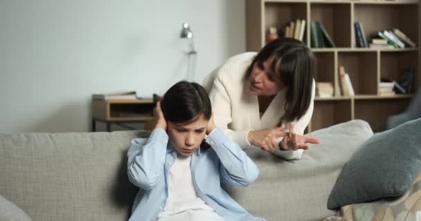 Стерн-кавказская мать продолжает ругать сына, который дерзко закрывает уши, чтобы блокировать выговор. Сцена изображает сложное родительское взаимодействие. - Кадры, видео