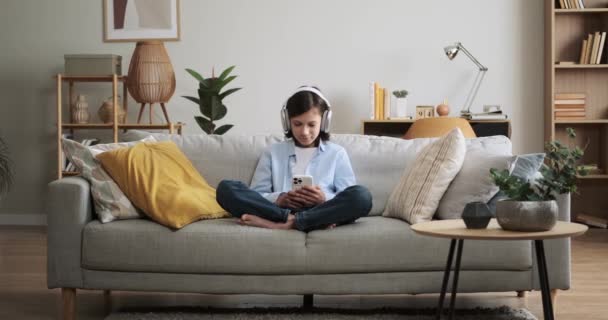 Young boy wearing headphones, immerses himself in the world of music while browsing content on phone. Seated comfortably on the living room couch, he finds solace in the melodies. - Footage, Video