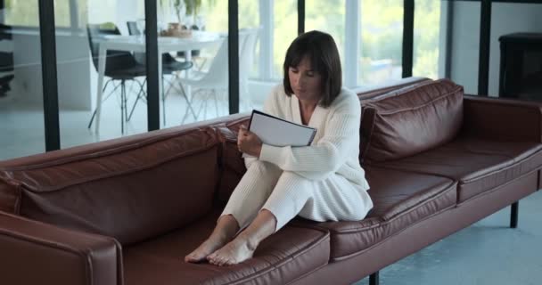 Keskittynyt valkoihoinen nainen istuu sohvalla, ahkerasti kirjoittaen muistikirjaan. Hänen huomaavainen ilme ja huolellinen muistiinpanot kuvaavat aura keskittymistä ja tuottavuutta. - Materiaali, video
